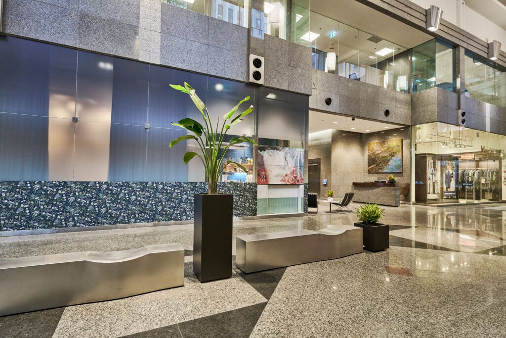 Modern commercial building lobby with indoor plants, polished floors, and retail storefronts.